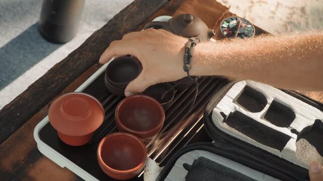 Beach tea ceremony with portable kit, clay teapot and cups arranged on wooden tray, hands opening textured travel case, leather strap and compact pouch revealed, steam rising from freshly poured tea,