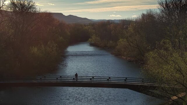 Drone video moving along the Boise River on an early spring morning, Idaho. The Friendship Bridge appears silhouetted against the soft morning light with peaceful riverside scenery.