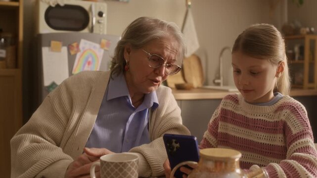 Tilt up shot of Caucasian granny asking her granddaughter about smartphone and social networks while they sitting at table in kitchen