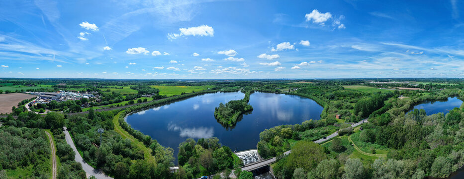 Aerial view of the expansive lakes and lush green landscapes of Kingsbury Water Park under a bright blue sky with fluffy clouds Kingsbury, England, United Kingdom.