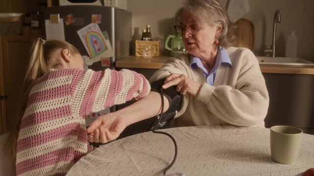 Handheld shot of Caucasian little girl helping her beloved granny with measuring blood pressure, using tonometer while they sitting at table