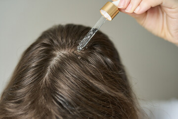 Fototapeta premium Woman Placing Clear Liquid Drops From Glass Pipette Onto Scalp Along Center Part Of Brown Hair In Closeup Indoor Grooming Scene Showing Precise Topical Application And Parted Roots.