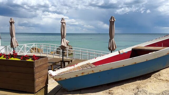 Coastal landscape with a colorful wooden boat on the sand, beach umbrellas, and a wooden deck overlooking the blue sea under a cloudy sky