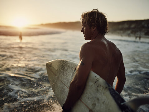 california surfer dude stands in ocean, holding surfboard as sun sets in background. warm light creates beautiful scene, highlighting waves and surfer focused