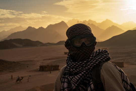 Epic mystery desert survivor profile portrait. 
Cinematic medium close up of man traveler in traditional 
scarf shemagh and dust goggles against golden mountains in Egypt.
