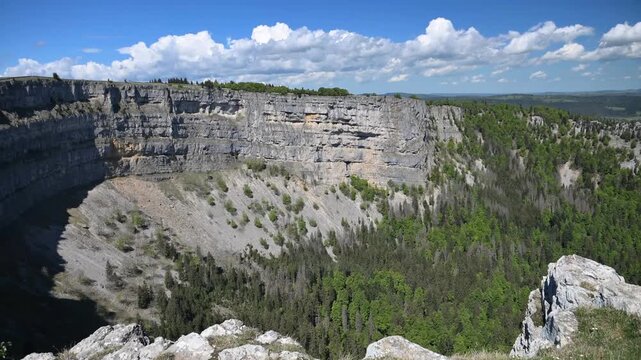 Panoramic view of the Creux du Van rocky amphitheater with green forest and dramatic clouds in springtime, Switzerland.