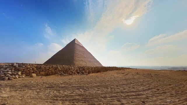 Cinematic timelapse of the Great Pyramid of Giza in Cairo, Egypt, with a camel rider briefly passing through the sandy desert foreground under a dramatic sky of moving clouds. The ancient stone wall r