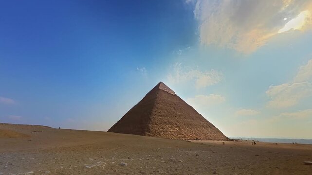 Dramatic timelapse video of the Great Pyramid of Giza in Cairo, Egypt, capturing the movement of clouds across a vivid blue sky, the slow arc of the sun, and tourists with camels passing through the s