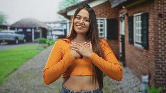 Woman with hands on chest smiling outside a brick building in an orange crop top and jeans; gratitude.