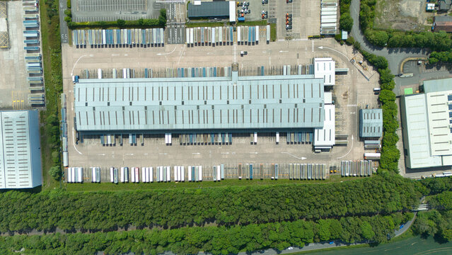 Aerial view of a large industrial warehouse with rows of parked semi-trailers and lush green trees in Kingsbury, England, United Kingdom.