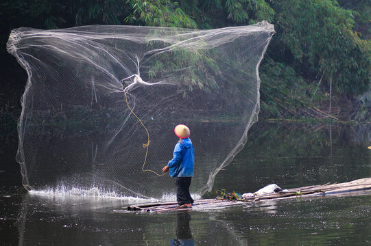 An Indonesian fisherman wearing a traditional conical hat stands on a bamboo raft, skillfully throwing a large fishing net over a river