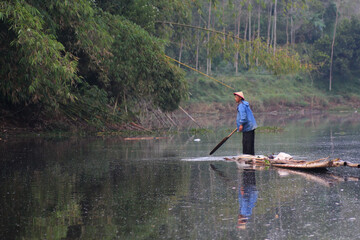 Naklejka premium Asian Indonesian male fisherman navigating a traditional bamboo raft on a calm river surrounded by lush nature.