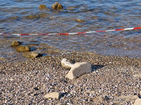 red and white plastic caution tape stretched across the frame. In the foreground, large concrete blocks and scattered river stones rest on a pebble beach. In the background, calm blue river water ripp