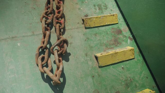 Rusted chain lying across green deck near yellow chocks, sunlight casting sharp shadows on worn paint, textured metal and oxidation stains, port maintenance scene with dockworker preparing load, quiet
