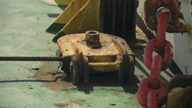Rusty yellow pulley on deck, detailed texture of corroded metal with red chain link and green painted surface, salt stains and flaking paint, mooring hardware under heavy load, tension cable