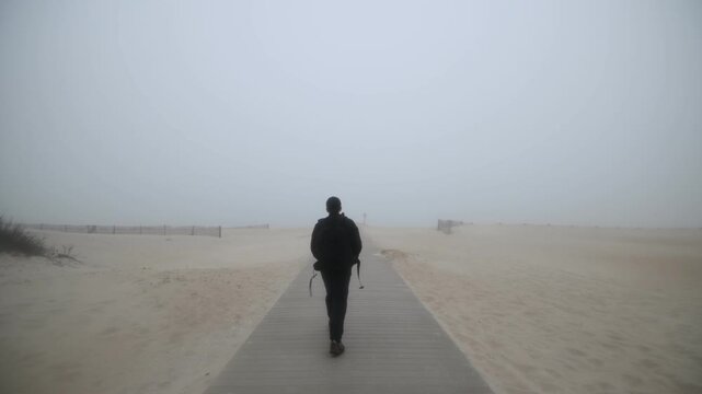 A hiker dressed in black and carrying a camera backpack walks along a wooden boardwalk toward a beach shrouded in dense fog at Assateague Island, Maryland. The scene conveys exploration, solitu