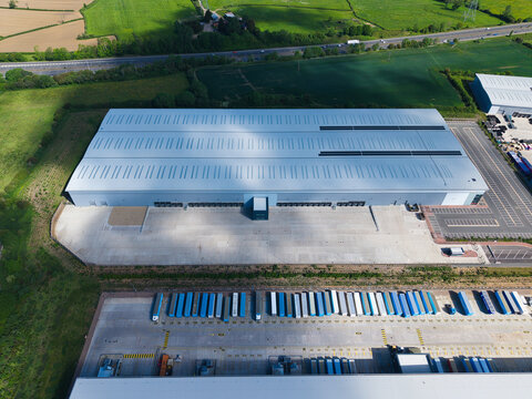 Aerial view of a large modern industrial warehouse with loading docks and rows of parked semi-trailers near green fields in Hinckley, England, United Kingdom.