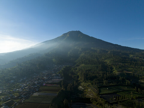 Aerial view of Mount Sumbing rising above the village of Garung and terraced agricultural fields under a clear blue sky in Kecamatan Kalikajar, Jawa Tengah, Indonesia.