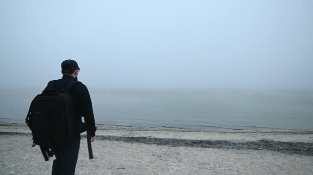 A photographer dressed in black with a camera backpack walks toward the beach at Assateague Island, Maryland, as dense sea fog rolls in from the ocean, creating a moody and atmospheric coastal scene