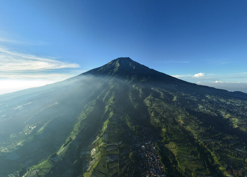 Aerial view of Mount Sumbing with terraced agricultural fields and a small village at the base under a clear blue sky in Kecamatan Kalikajar, Jawa Tengah, Indonesia.