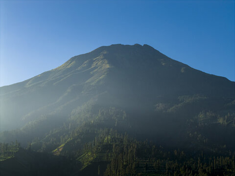 Aerial view of Mount Sumbing and the Garung area featuring terraced fields and misty slopes under a clear blue sky in Kecamatan Kalikajar, Jawa Tengah, Indonesia.