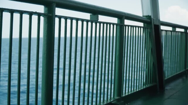 Ferry railing overlooking calm blue ocean, rusted green metal balustrade framing steady horizon, gentle waves lapping, soft daylight, cinematic minimal composition, deck planks and safety post,