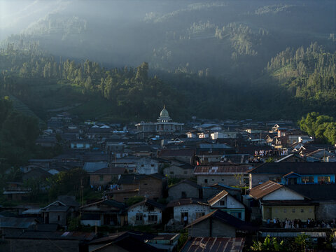 Aerial view of the village of Garung with people gathered near a golden-domed mosque as morning light filters through misty green hills in Kecamatan Kalikajar, Jawa Tengah, Indonesia.