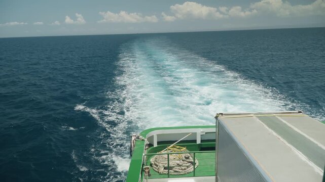 Green deck ferry leaving foamy wake, open ocean transit with turquoise trail and deck equipment, clear horizon and scattered clouds, brisk motion and coastal route energy, commercial shipping vibe
