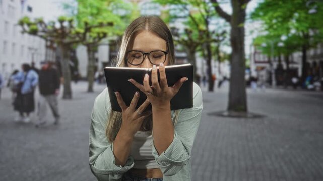Woman holding book close to face with hands on pages on a cobblestone street; curiosity study leisure.