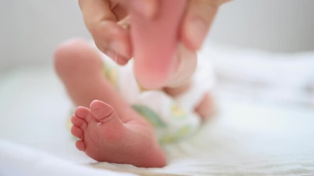 Close up view of a mother's hand gently caressing her newborn baby's tiny feet. The infant, wearing a diaper, lies on a soft white surface, experiencing a moment of loving connection