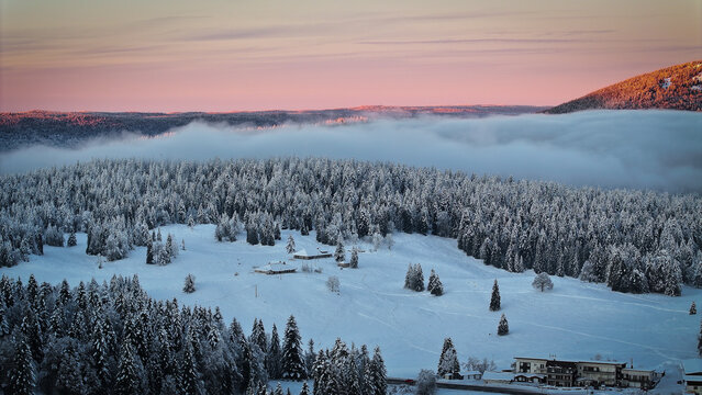 Aerial view of the snowy landscape of Les Rousses with dense pine forests, rolling fog, and a soft pink sunset sky in Les Rousses, Bourgogne-Franche-Comte, France.