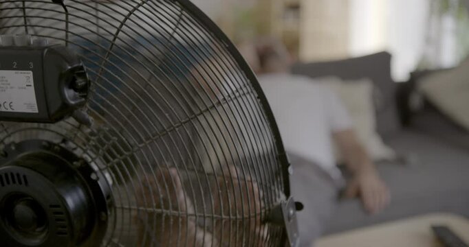 Cool Breeze Relief: A person finds comfort in the cooling breeze of an electric fan, seeking respite from the summer heat indoors.