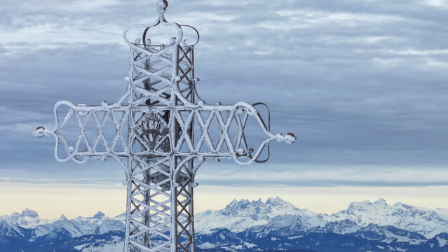 Aerial view of the frost-covered Le Reculet summit cross with snow-capped Alpine peaks under a cloudy sky Thoiry, Auvergne-Rhone-Alpes, France.
