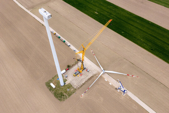 Aerial view of a wind turbine under construction with a large yellow crane and blades on the ground amidst agricultural fields in Zielona, Masovian Voivodeship, Poland.