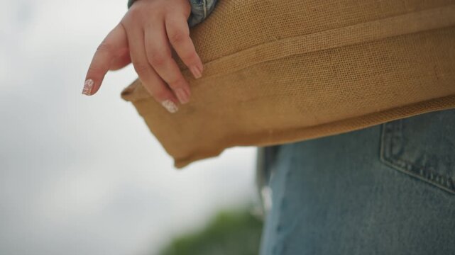 Close up hand resting on woven tote, denim sleeve, cloudy sky backdrop, delicate manicure, slow motion gesture, contemplative mood, sustainable texture, commuter pausing between errands on quiet
