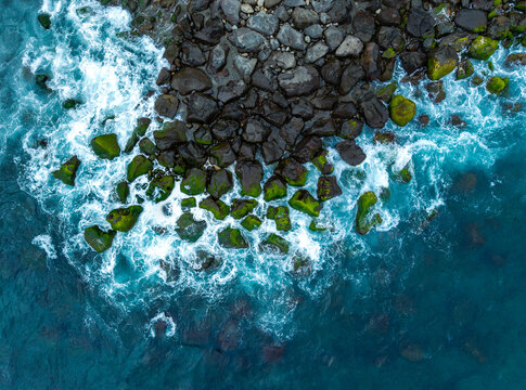 Aerial view of crashing white waves against dark volcanic rocks covered in green moss along the turquoise coastline of Povoacao, Acores, Portugal.