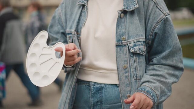 Artist holding paint palette outdoors, denim jacket and jeans, closeup of hands gripping white plastic palette with empty wells, casual street park backdrop, soft natural light, candid creative moment