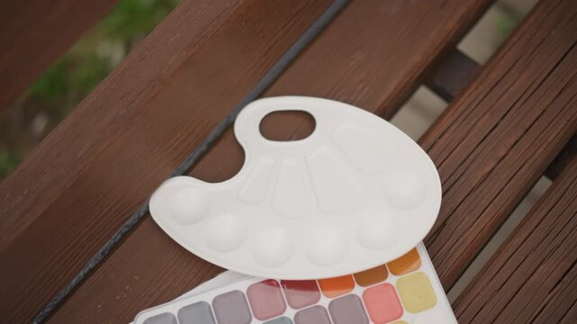 White palette watercolor set on bench, artist placing empty plastic palette beside pastel swatch sheet on slatted wood bench, soft natural sunlight, muted color tones, quiet creative moment