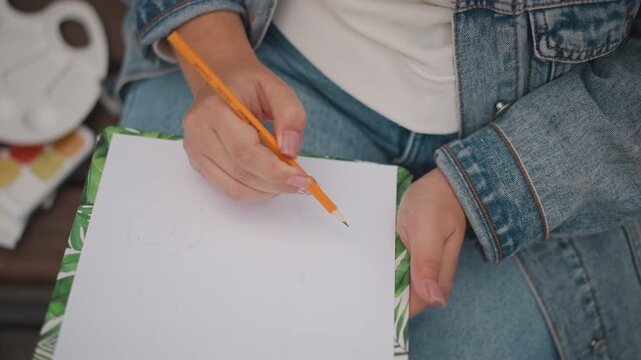 Hands sketching on blank sheet with pencil, denim jacket on outdoor bench, paint palette visible, relaxed art session, natural light and soft bokeh, hobbyist practice and inspiration