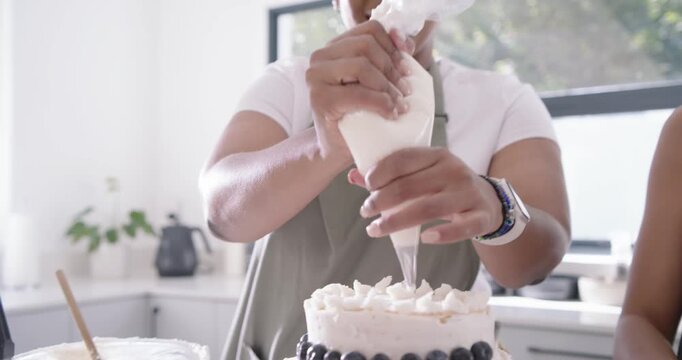 Diverse female friends piping from bag and placing decor on two-tier cake in kitchen for party