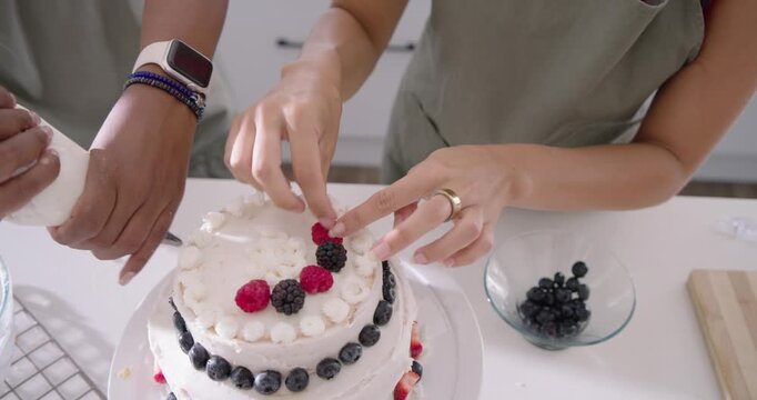 Diverse women in green aprons near finished cake, placing berries and piping frosting at counter