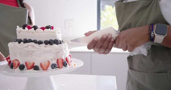 Diverse female bakers finishing cake piping bag turning cake on kitchen counter adding berries