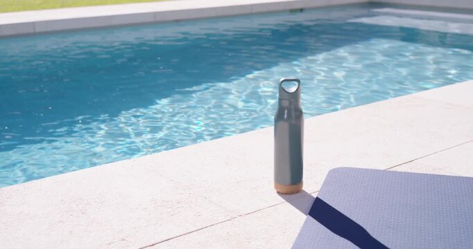 Metal water bottle is standing on pool deck while camera is pulling back, revealing sparkling pool