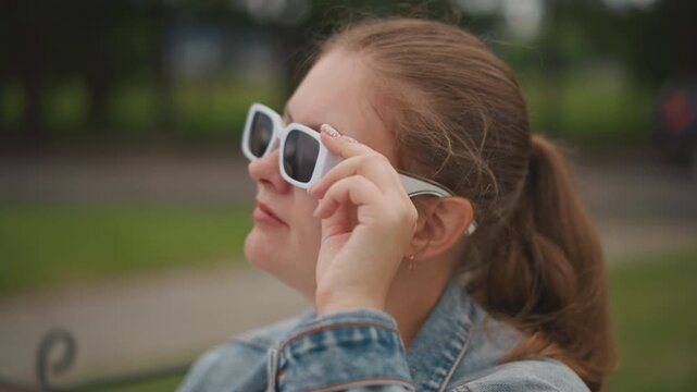 White woman adjusts sunglasses on bench with confident smile, denim jacket, ponytail, playful vintage frames, warm park greenery, shallow depth, influencer energy and street style vibe as she tests