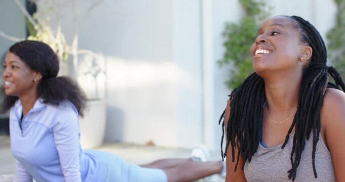 African women friends in sportswear smiling, practicing cobra on deck on green mats with bottle
