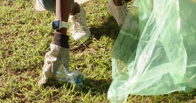 African American woman spotting bottle on grass squatting placing bottle into green bag for cleanup