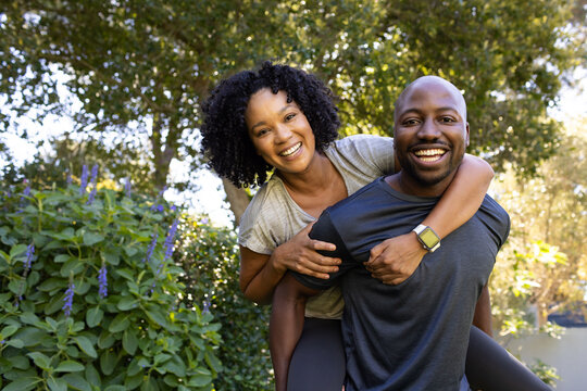 African American couple smiling man carrying woman on back in garden in sportswear, smartwatch