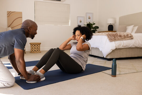 African American couple in gymwear, woman crunching on mat at home, water bottle, man holding feet