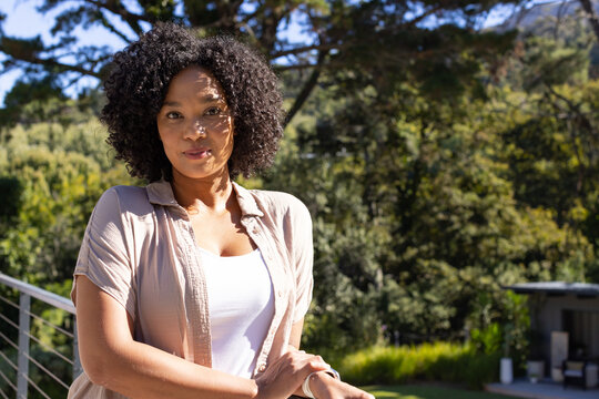 African American woman standing on balcony by cable railing in garden wearing beige shirt, bracelet