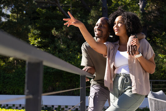 African American couple standing on raised deck pointing left by railing, woman wearing smartwatch